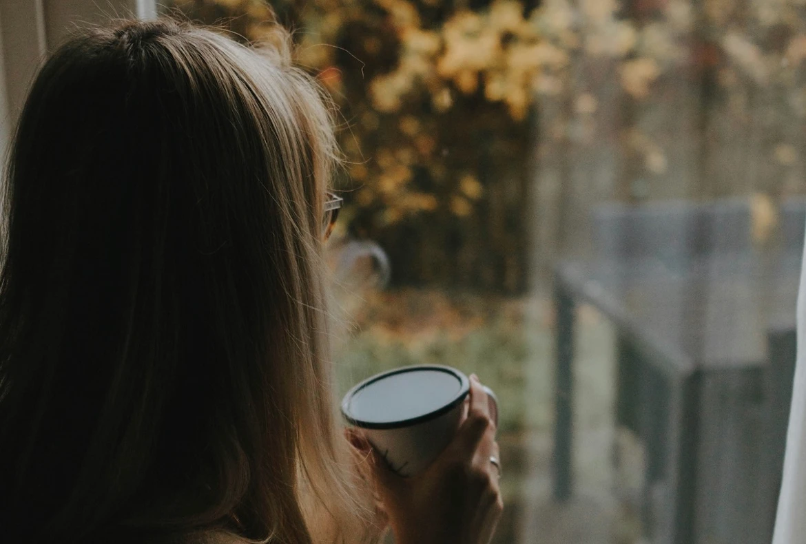 Mujer mirando por la ventana con una taza de café, simbolizando la reflexión y la culpa de no llegar a todo.